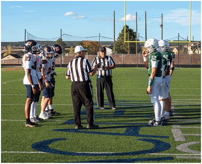 People are gathered on a sunlit football field with goalposts and a 50-yard marker visible under a partly cloudy sky. Description generated by AI