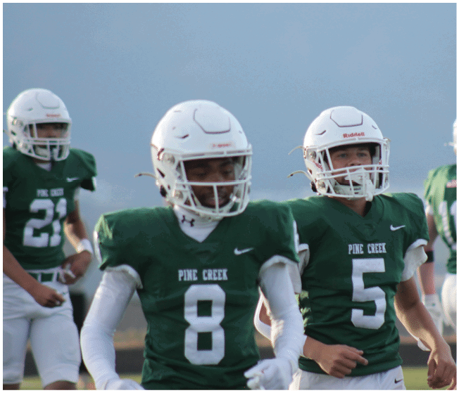 Three people wearing green football uniforms and white helmets are on a field, with two facing forward and one facing away. Description generated by AI