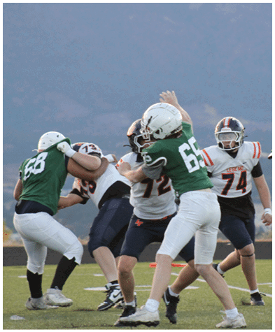 Two players in green jerseys tackle a player in a white jersey during a football game while a person in the background records the action with a camera on a tripod. Description generated by AI