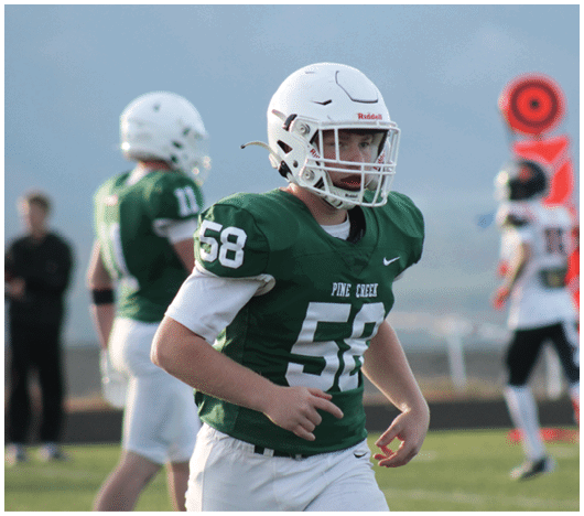 A person wearing a green football jersey and white helmet is running on a field with other players and a blurred figure in the background. Description generated by AI