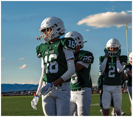 Several people wearing green and white football uniforms and helmets stand on a field under a clear blue sky with mountains in the background. Description generated by AI