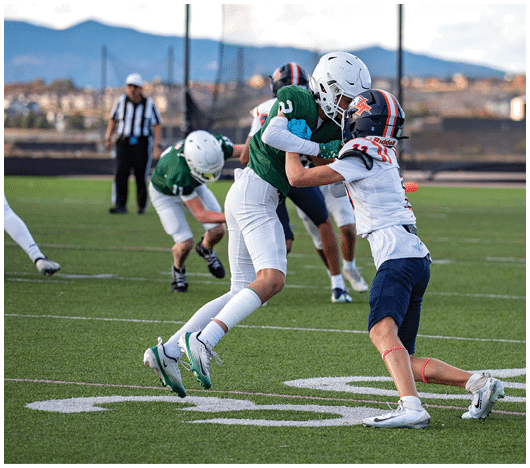 Two people in football uniforms are engaged in a tackle on a green field with a referee and other players in the background. Description generated by AI