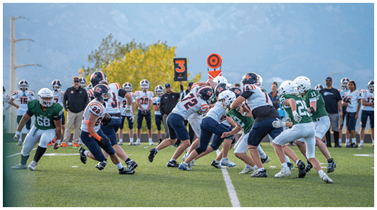 A group of people wearing football uniforms are engaged in a play on a grassy field with others standing on the sidelines and a mountainous background. Description generated by AI