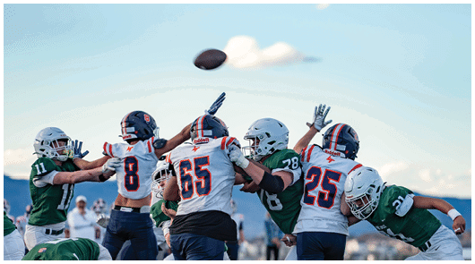People wearing football uniforms are actively reaching and jumping to catch a football in mid-air during a game. Description generated by AI