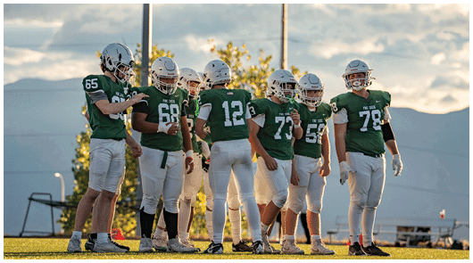 A group of people wearing green football jerseys and helmets are standing together on a field with a cloudy sky and mountains in the background. Description generated by AI
