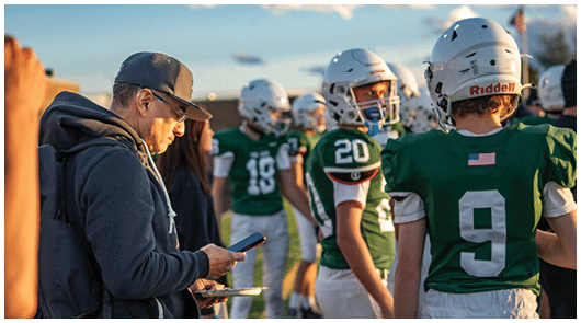 A person wearing a cap and backpack is looking at a phone while standing near several people in green football uniforms on a field during sunset. Description generated by AI