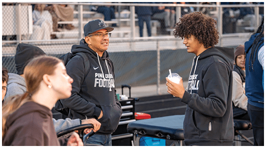 Two people wearing hoodies with “Pine Creek Football" printed on them are engaged in conversation near sports equipment and bleachers. Description generated by AI