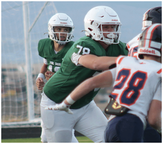 Two people in green football uniforms and white helmets are engaged in a play against two people in white and blue football uniforms with orange accents on a field. Description generated by AI