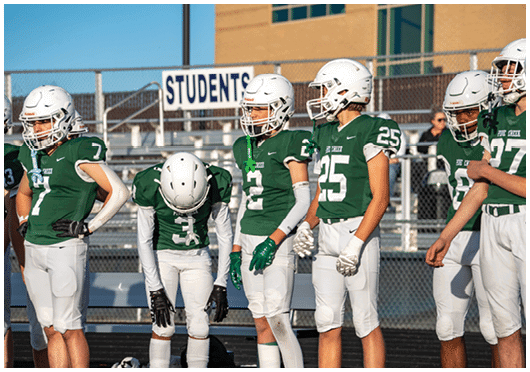 Four people in green and white football uniforms stand on a field with one person bent over and the others standing upright near bleachers and a sign that reads “STUDENTS." Description generated by AI