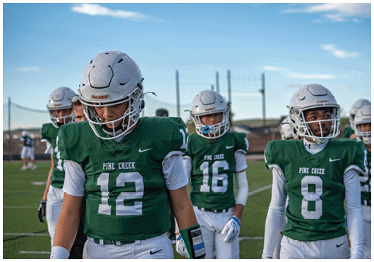 Several people wearing green football uniforms and helmets stand on a field with a clear sky in the background. Description generated by AI