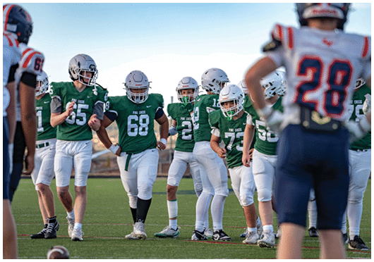 A group of people wearing green and white football uniforms are standing on a field while another person in a blue and white uniform with the number 28 is in the foreground facing them. Description generated by AI