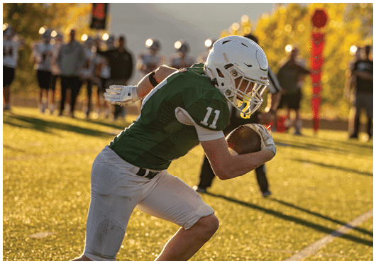 A person wearing a green football jersey and white helmet is running on a sunlit field while holding a football tightly. Description generated by AI