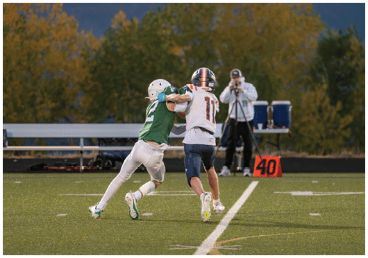Two people in football uniforms are engaged in a tackle on a field with a person in the background filming the action near a marker showing the number 40. Description generated by AI