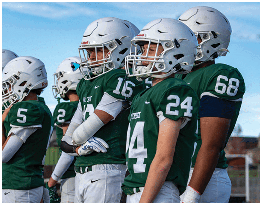 People wearing green football uniforms and white helmets stand in a row outdoors under a blue sky. Description generated by AI