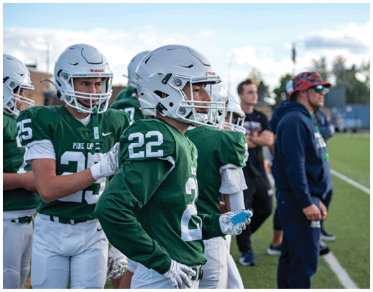 People in green football uniforms and helmets stand on the sidelines of a football field while others in casual clothing stand nearby. Description generated by AI