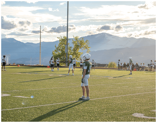 Several people wearing football uniforms and helmets are standing on a grassy field with mountains and a partly cloudy sky in the background. Description generated by AI
