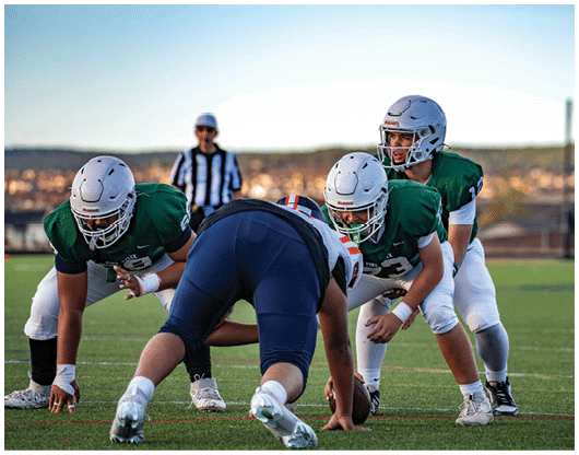 Three football players in green jerseys and white helmets are positioned on the field facing a player in a dark uniform with a helmet, while a referee stands in the background. Description generated by AI