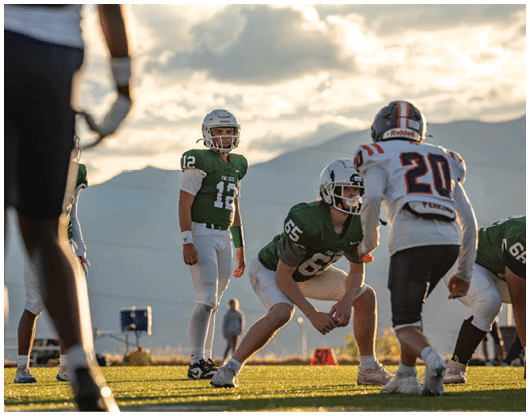 Several people in football uniforms are positioned on a field at sunset, preparing for a play with mountains visible in the background. Description generated by AI
