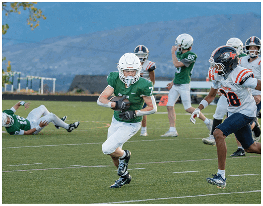A person in a green football uniform is running with the ball on a field while players in white and blue uniforms pursue and one player in green is falling on the ground. Description generated by AI