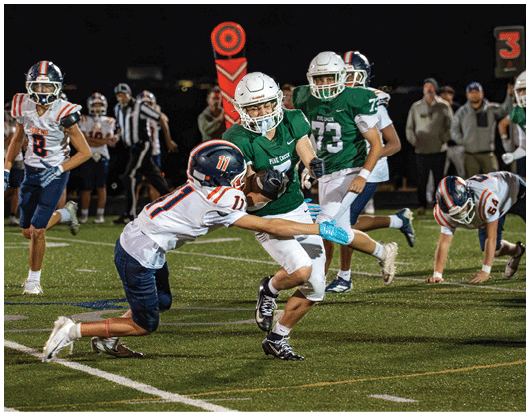 People wearing football uniforms are actively playing on a field at night with one person in green running while another in white and blue attempts to tackle. Description generated by AI