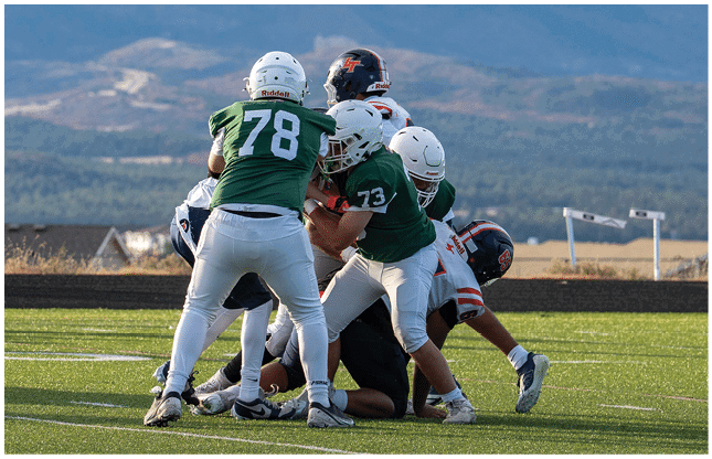 Several people wearing football helmets and uniforms are engaged in a tackle on a grassy field with mountains in the background. Description generated by AI