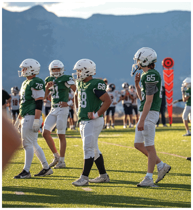Four people wearing green football jerseys and white helmets stand on a sunlit field with other players and spectators in the background. Description generated by AI