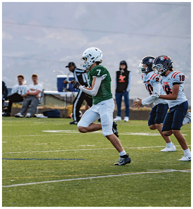 A person wearing a green football jersey and white helmet is running on a football field with spectators and a referee in the background. Description generated by AI