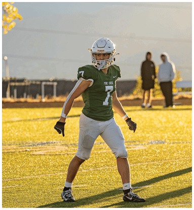 A person wearing a green football jersey with the number seven and white pants stands on a sunlit football field with two people in the background. Description generated by AI
