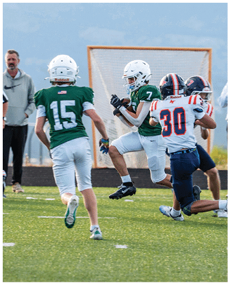 Two people in green football uniforms are running on a field while two people in white and blue football uniforms attempt to block them near a goal net. Description generated by AI