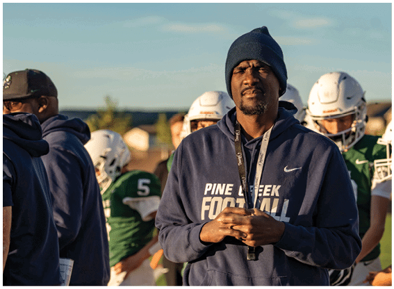 A person wearing a navy blue hoodie with “Pine Creek Football" printed on it and a dark beanie stands with hands clasped while several people in football helmets and green jerseys are in the background. Description generated by AI