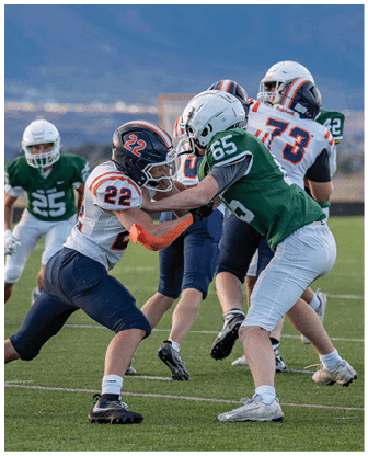 Two people in football uniforms are engaged in a play on a field while a third person in a green uniform stands in the background. Description generated by AI