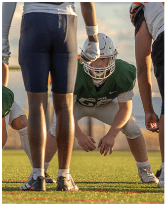 People in football uniforms are positioned on a field, with one person in a green jersey crouched and ready to play. Description generated by AI