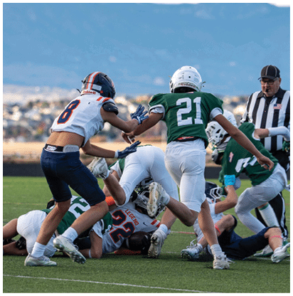 Several people wearing football uniforms are engaged in a play on a grassy field with a blurred background of buildings and mountains. Description generated by AI