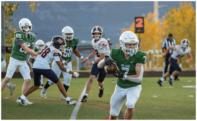 People in football uniforms are playing on a field with one person in a green jersey running with the ball while others in white and dark jerseys are chasing. Description generated by AI