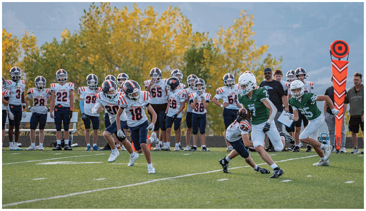 Children in football uniforms are playing on a field with some standing on the sidelines and others actively running and tackling during a game. Description generated by AI
