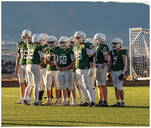A group of people in green and white football uniforms stand together on a field while one person in a white and red uniform stands apart facing them with mountains in the background. Description generated by AI