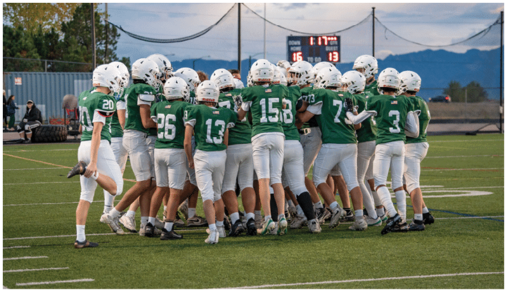 A group of people wearing green football uniforms and white helmets are huddled together on a football field with a scoreboard and mountains visible in the background. Description generated by AI