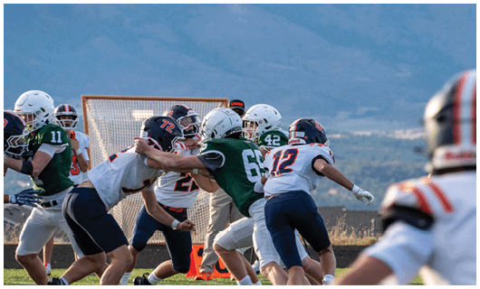 People wearing helmets and football uniforms are engaged in a play on a field with a goalpost and mountains in the background. Description generated by AI