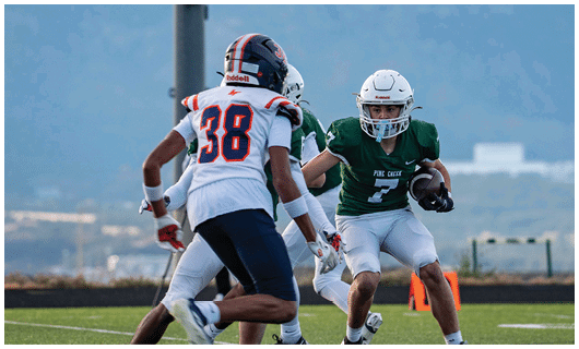 A person in a green football uniform is running with the ball while two people in white and navy uniforms attempt to block or tackle on a football field. Description generated by AI