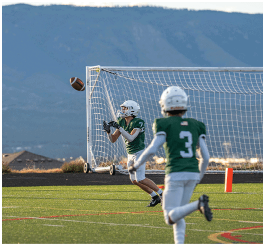 Two people in green sports uniforms and helmets are playing football near a goalpost on a field with mountains in the background. Description generated by AI
