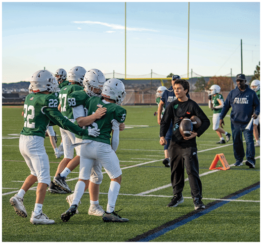 People in football uniforms are celebrating on a field while another person in casual clothing holds a football and smiles nearby. Description generated by AI