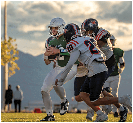 A group of people wearing football uniforms are actively playing on a field with mountains and a cloudy sky in the background. Description generated by AI