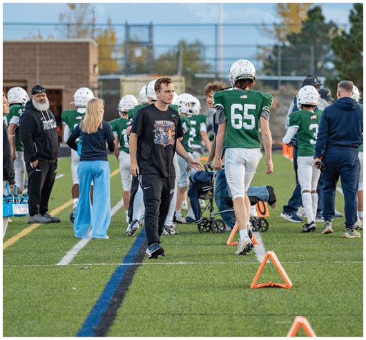 People in sports uniforms and casual clothing are gathered on a football field near the sideline with cones marking the area. Description generated by AI