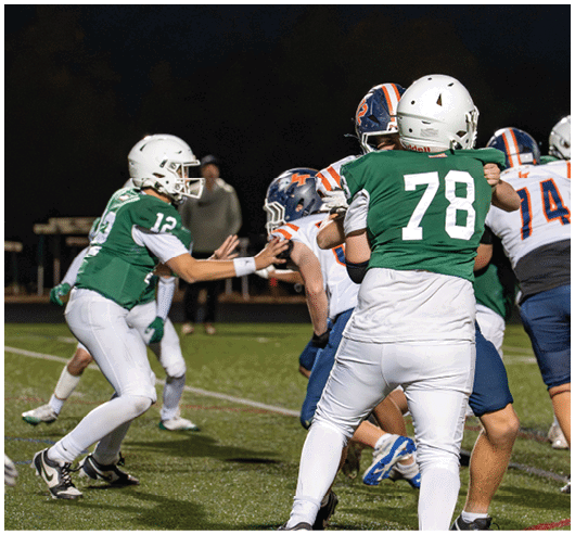 Players in green and white uniforms are engaged in a nighttime American football game against players in white and blue uniforms on a grassy field. Description generated by AI