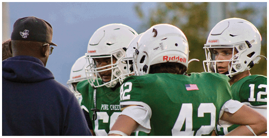 Two people in green football uniforms with white numbers and one person in a dark jacket are standing close together, with one football player holding the other's arm. Description generated by AI
