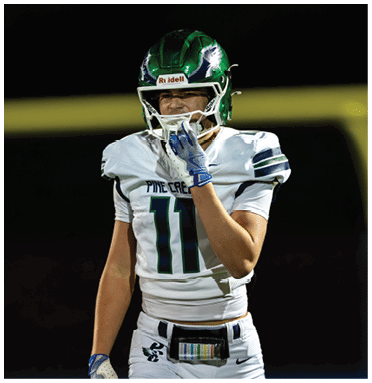 A person wearing a green and white football helmet and a white football uniform with the number 11 is standing on a field at night, holding their chin strap with a gloved hand. Description generated by AI