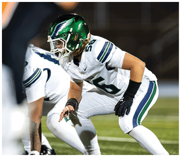 Two people in football uniforms and helmets are crouched in a ready position on a field during a game. Description generated by AI