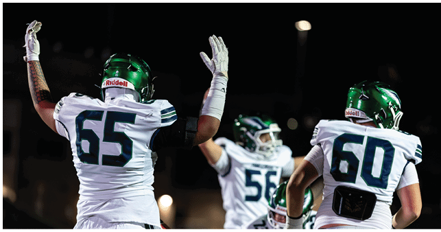 Three football players in white uniforms and green helmets are raising their arms on a field at night. Description generated by AI