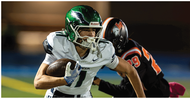 A person wearing a green helmet and white football uniform is running with a football while another person in a black and orange uniform is behind them. Description generated by AI
