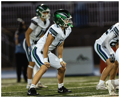 Two people in football uniforms and helmets are positioned on a field, appearing ready for action. Description generated by AI